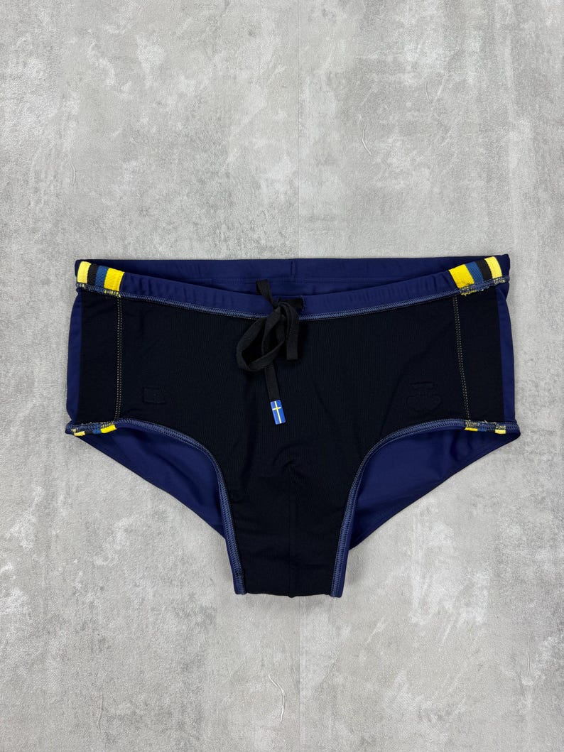 May include: Black and navy blue swim briefs with yellow and blue accents on the waistband. The briefs have a drawstring closure and a small logo detail. The swimwear is designed for swimming and water activities.