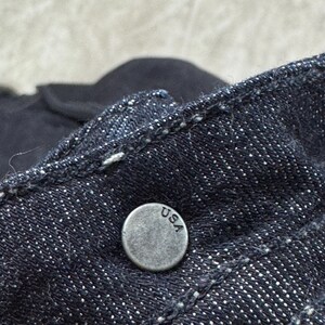 Puede incluir: Una tela de mezclilla azul &iacute;ndigo oscuro con un bot&oacute;n met&aacute;lico circular de acabado plateado desgastado. El bot&oacute;n tiene grabadas las letras USA en una fuente sencilla sin remates. El material de mezclilla muestra un tejido de sarga y costuras blancas.