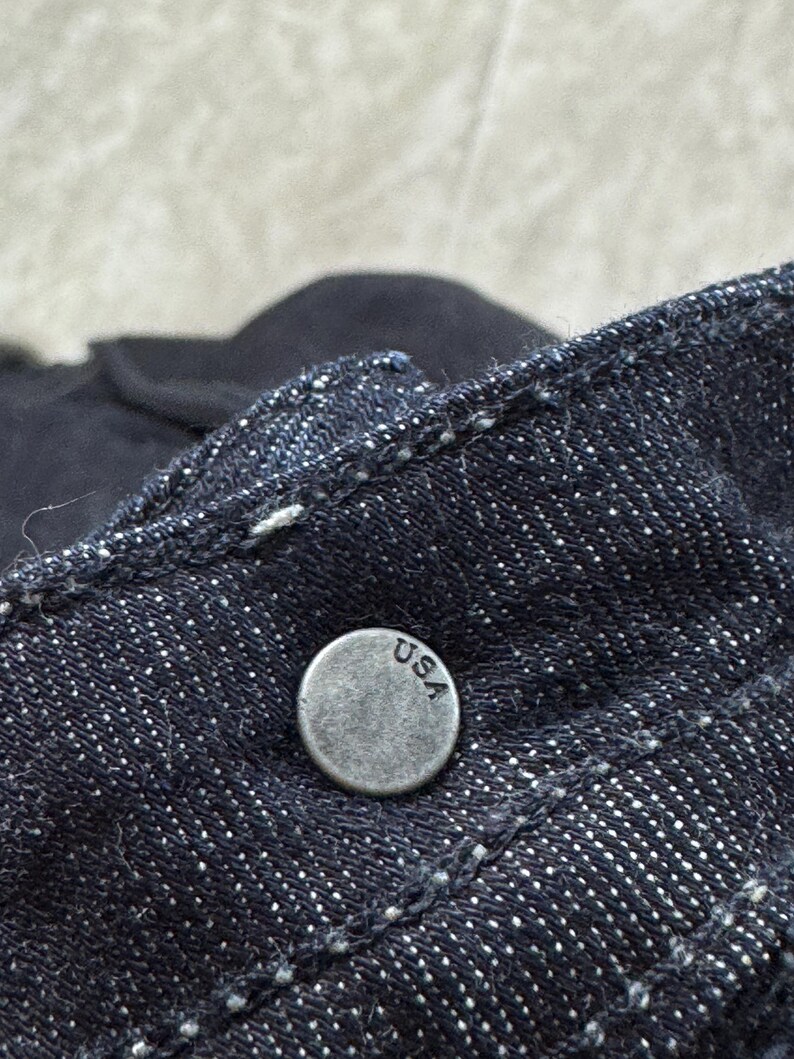 Puede incluir: Una tela de mezclilla azul &iacute;ndigo oscuro con un bot&oacute;n met&aacute;lico circular de acabado plateado desgastado. El bot&oacute;n tiene grabadas las letras USA en una fuente sencilla sin remates. El material de mezclilla muestra un tejido de sarga y costuras blancas.
