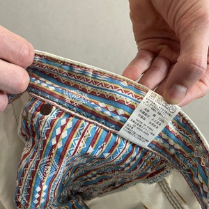 May include: A white pair of jeans with a patterned waistband. The waistband is made of a cotton fabric with a blue, red, and white geometric pattern. The label on the waistband reads "本体 綿 100% 付属部分 人工皮革 企画生産㈱エドウイン商事 MADE IN CHINA 03-5604-8900 ▲注意 汗や雨などで湿った状態での摩擦により色移りすることがあります。"