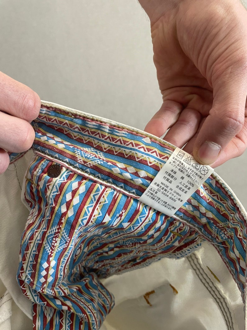 May include: A white pair of jeans with a patterned waistband. The waistband is made of a cotton fabric with a blue, red, and white geometric pattern. The label on the waistband reads "本体 綿 100% 付属部分 人工皮革 企画生産㈱エドウイン商事 MADE IN CHINA 03-5604-8900 ▲注意 汗や雨などで湿った状態での摩擦により色移りすることがあります。"