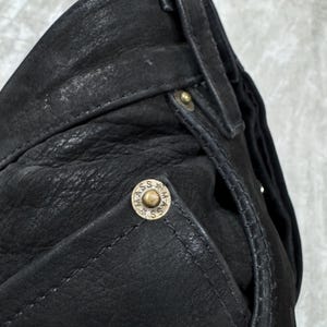 May include: A pair of black leather trousers featuring a textured surface, belt loops, and a metallic rivet on the pocket corner. The rivet is circular, brass-toned, and embossed with the text MASS along with small star symbols around the centre.