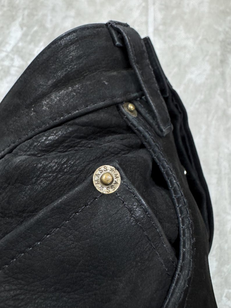 May include: A pair of black leather trousers featuring a textured surface, belt loops, and a metallic rivet on the pocket corner. The rivet is circular, brass-toned, and embossed with the text MASS along with small star symbols around the centre.