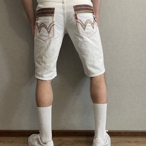 May include: White denim shorts with embroidered details on the back pockets. The shorts have a distressed hem and are worn with white socks and white sneakers.
