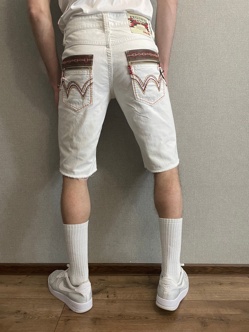 May include: White denim shorts with embroidered details on the back pockets. The shorts have a distressed hem and are worn with white socks and white sneakers.