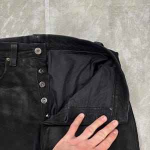 May include: Black leather trousers featuring a button-fly closure with four visible silver-toned metal buttons and a matching top button at the waistband. The interior is lined with black fabric, and the waistband includes belt loops.