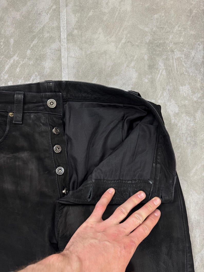 May include: Black leather trousers featuring a button-fly closure with four visible silver-toned metal buttons and a matching top button at the waistband. The interior is lined with black fabric, and the waistband includes belt loops.