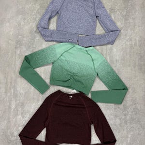 May include: Three long-sleeved crop tops in shades of purple, green, and burgundy. The tops have a textured knit design and thumb holes. The brand logo is visible on the back of each top. The tops are designed for athletic wear.