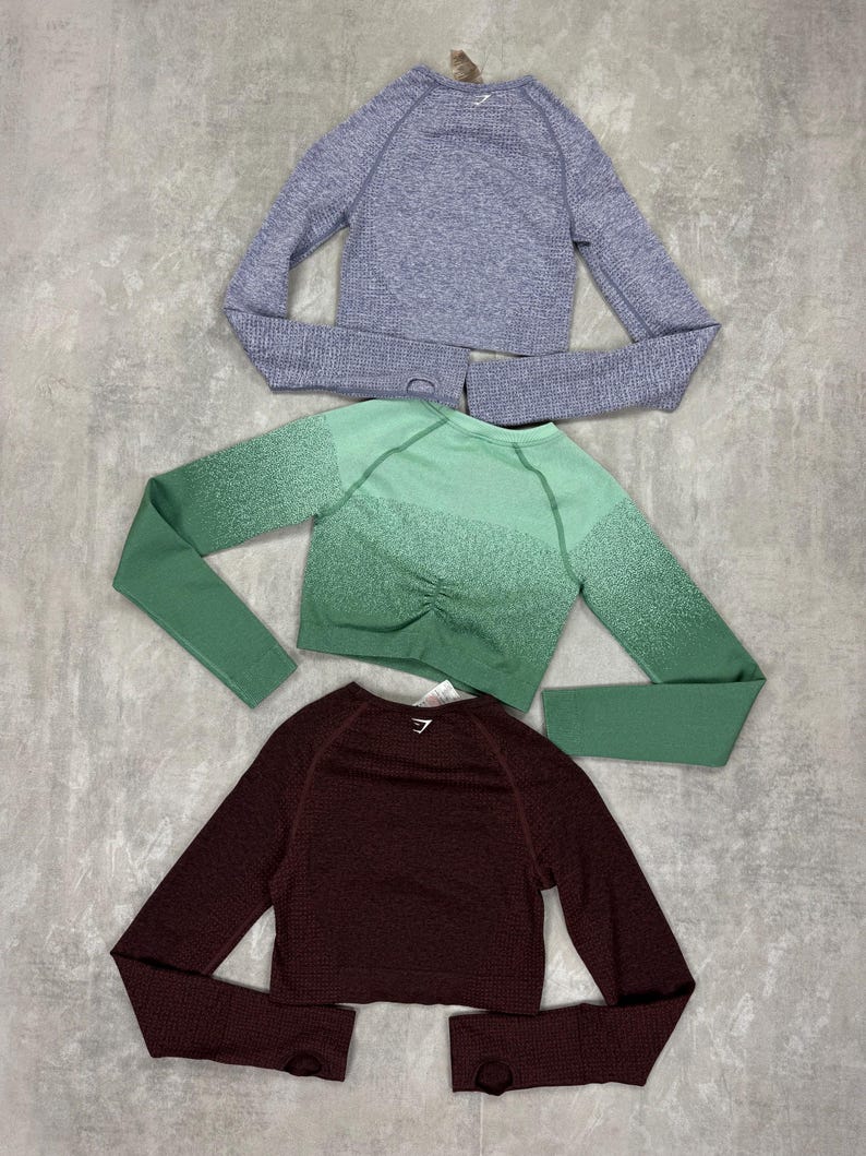 May include: Three long-sleeved crop tops in shades of purple, green, and burgundy. The tops have a textured knit design and thumb holes. The brand logo is visible on the back of each top. The tops are designed for athletic wear.
