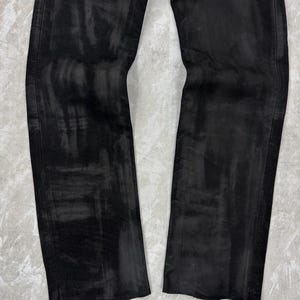 May include: A pair of black trousers featuring a distressed, washed-out finish with visible brushstroke-like patterns across the fabric. The trousers have a straight-leg cut with vertical side seams and a textured appearance throughout the material.
