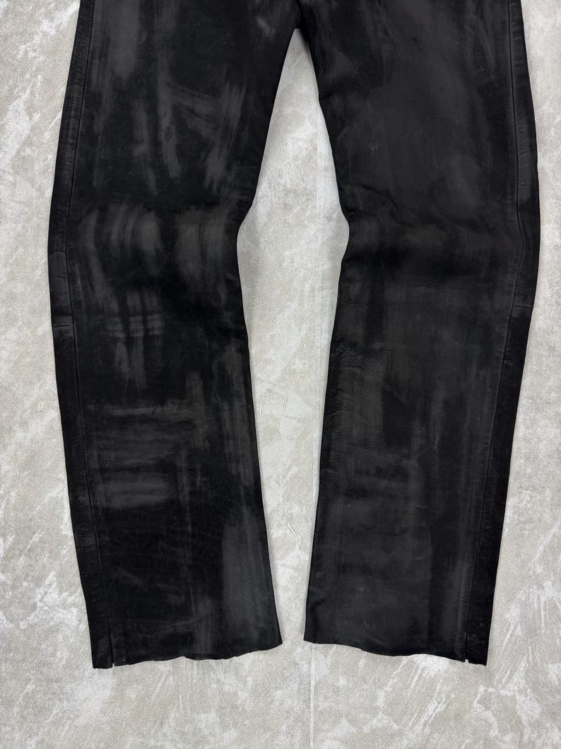 May include: A pair of black trousers featuring a distressed, washed-out finish with visible brushstroke-like patterns across the fabric. The trousers have a straight-leg cut with vertical side seams and a textured appearance throughout the material.