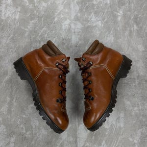 Puede incluir: Un par de botas de senderismo de cuero marrón con cordones marrones oscuros y suela negra. Las botas tienen un cuello acolchado y están diseñadas para actividades al aire libre. Las botas se muestran sobre un fondo gris.