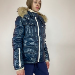Puede incluir: Una chaqueta acolchada azul marino con una cremallera blanca y ribetes blancos. La chaqueta tiene una capucha de piel sint&eacute;tica y varios bolsillos.