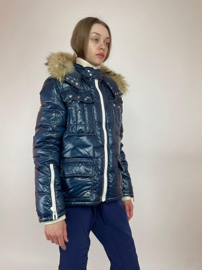 Puede incluir: Una chaqueta acolchada azul marino con una cremallera blanca y ribetes blancos. La chaqueta tiene una capucha de piel sint&eacute;tica y varios bolsillos.