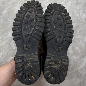May include: A pair of worn, brown leather work boots, soles facing up. The black rubber soles have a deep tread pattern and the CAT logo. The boots show signs of wear and use, with dirt and debris visible on the soles.