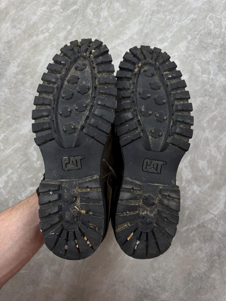 May include: A pair of worn, brown leather work boots, soles facing up. The black rubber soles have a deep tread pattern and the CAT logo. The boots show signs of wear and use, with dirt and debris visible on the soles.