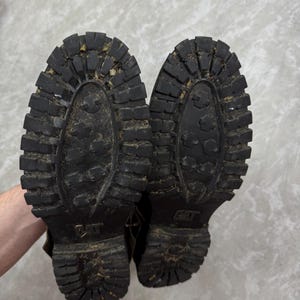 May include: A pair of worn black Caterpillar work boots, soles facing up. The soles have a deep tread pattern and the Caterpillar logo is visible. The boots show signs of wear and use, with dirt and debris in the treads.