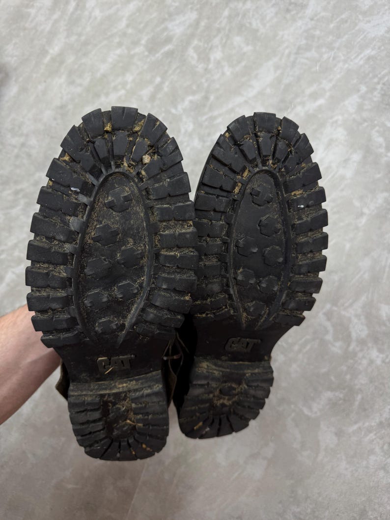 May include: A pair of worn black Caterpillar work boots, soles facing up. The soles have a deep tread pattern and the Caterpillar logo is visible. The boots show signs of wear and use, with dirt and debris in the treads.