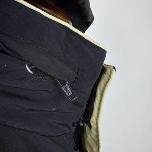 Puede incluir: Primer plano de una chaqueta con capucha negra con ribete beige. La chaqueta tiene cierre de cremallera y un logotipo en el tirador de la cremallera. La capucha est&aacute; doblada hacia atr&aacute;s, revelando el forro beige.