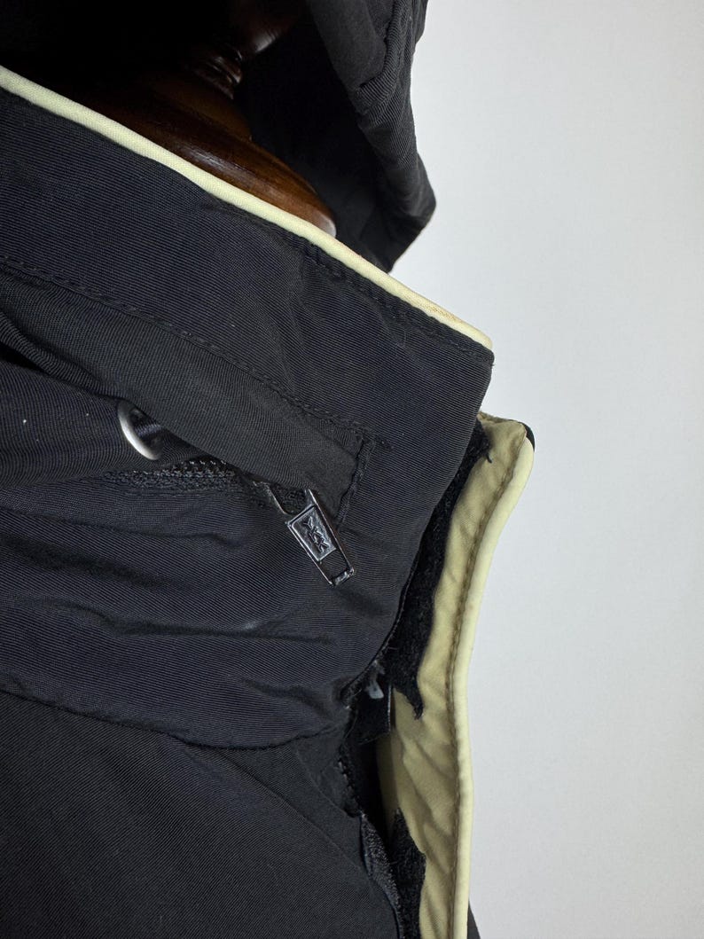 Puede incluir: Primer plano de una chaqueta con capucha negra con ribete beige. La chaqueta tiene cierre de cremallera y un logotipo en el tirador de la cremallera. La capucha est&aacute; doblada hacia atr&aacute;s, revelando el forro beige.