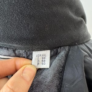 Puede incluir: Primer plano de un cuello de chaqueta negro con un forro polar suave y texturizado. Una etiqueta muestra informaci&oacute;n de tallas: USA XXL, I 58, D 58, F 50. El forro interior de la chaqueta es visible, con un patr&oacute;n sutil.