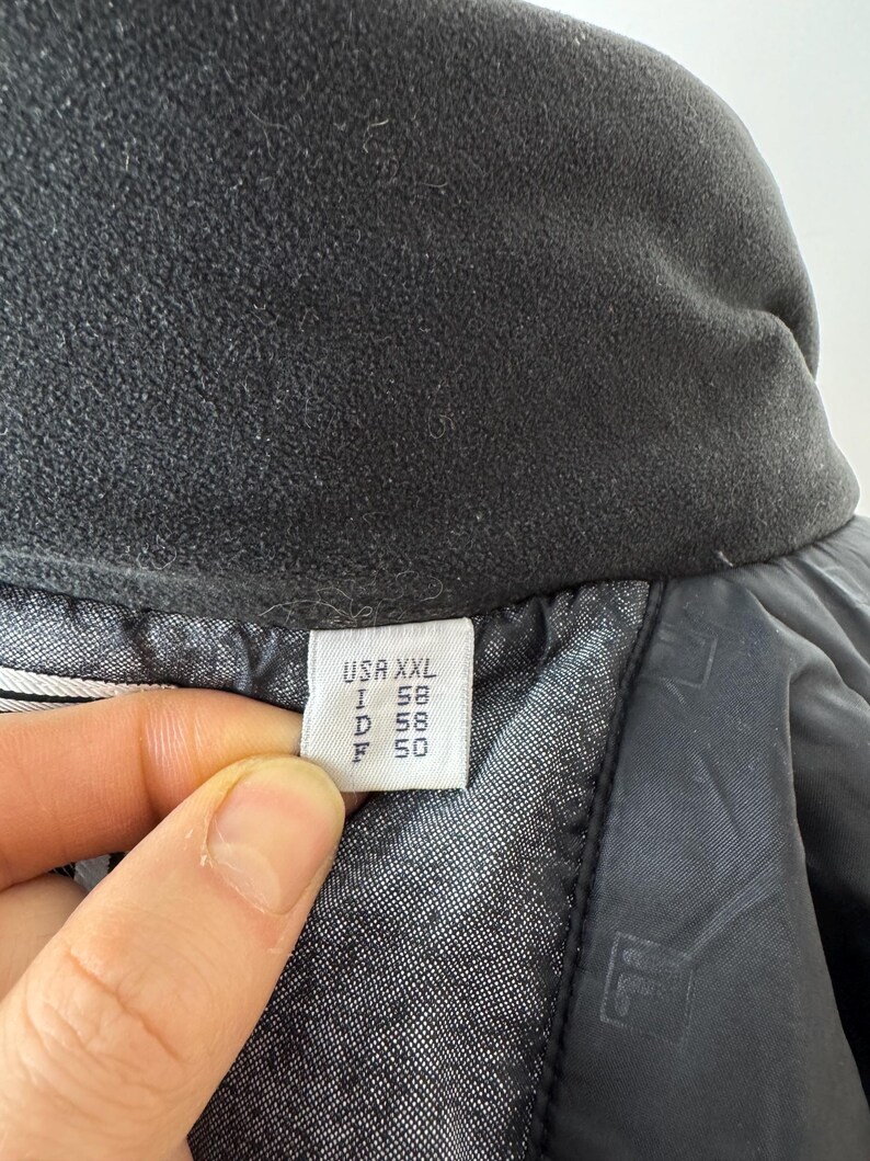 Puede incluir: Primer plano de un cuello de chaqueta negro con un forro polar suave y texturizado. Una etiqueta muestra informaci&oacute;n de tallas: USA XXL, I 58, D 58, F 50. El forro interior de la chaqueta es visible, con un patr&oacute;n sutil.