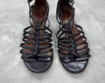 Puede incluir: Un par de sandalias de cuero negro con múltiples correas y un detalle trenzado en la parte delantera. Las sandalias tienen un cierre de hebilla y una plantilla marrón. Los zapatos se exhiben sobre una superficie gris.