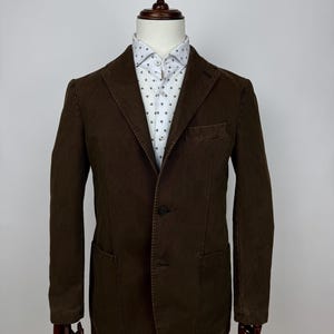 Può includere: Un blazer marrone con chiusura a due bottoni, due tasche frontali e una tasca sul petto. Il blazer è esposto su un manichino e abbinato a una camicia bianca button-down con un piccolo motivo ripetuto. Il colore del blazer è un marrone intenso e terroso.