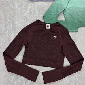 May include: A maroon long-sleeve crop top with a textured pattern and the brand logo on the chest. The top has a crew neck and is displayed with other athletic apparel in shades of green and gray.