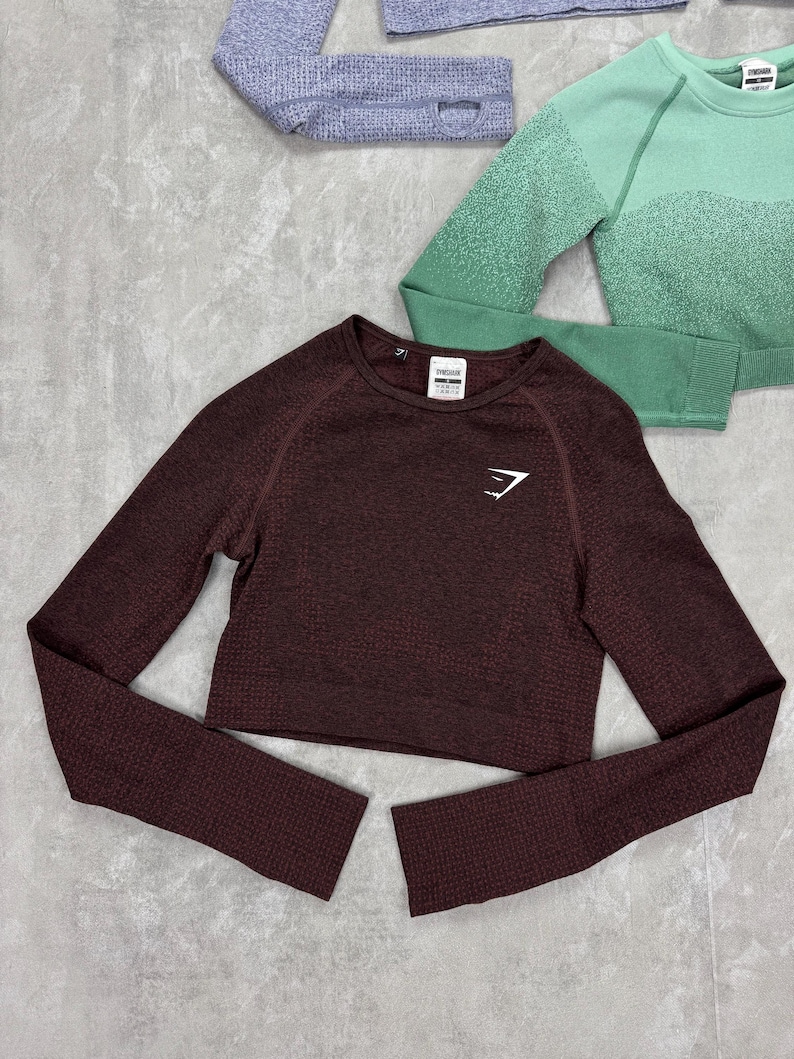 May include: A maroon long-sleeve crop top with a textured pattern and the brand logo on the chest. The top has a crew neck and is displayed with other athletic apparel in shades of green and gray.