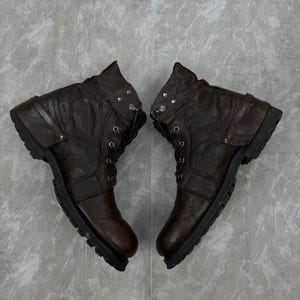 May include: A pair of brown leather boots with a rugged, worn appearance. The boots have a lace-up design, a rounded toe, and a thick, textured sole. The ankle area features decorative metal studs and straps.