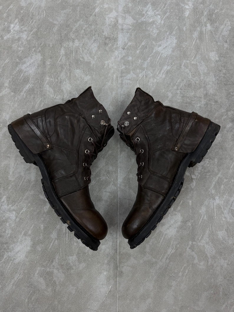 May include: A pair of brown leather boots with a rugged, worn appearance. The boots have a lace-up design, a rounded toe, and a thick, textured sole. The ankle area features decorative metal studs and straps.