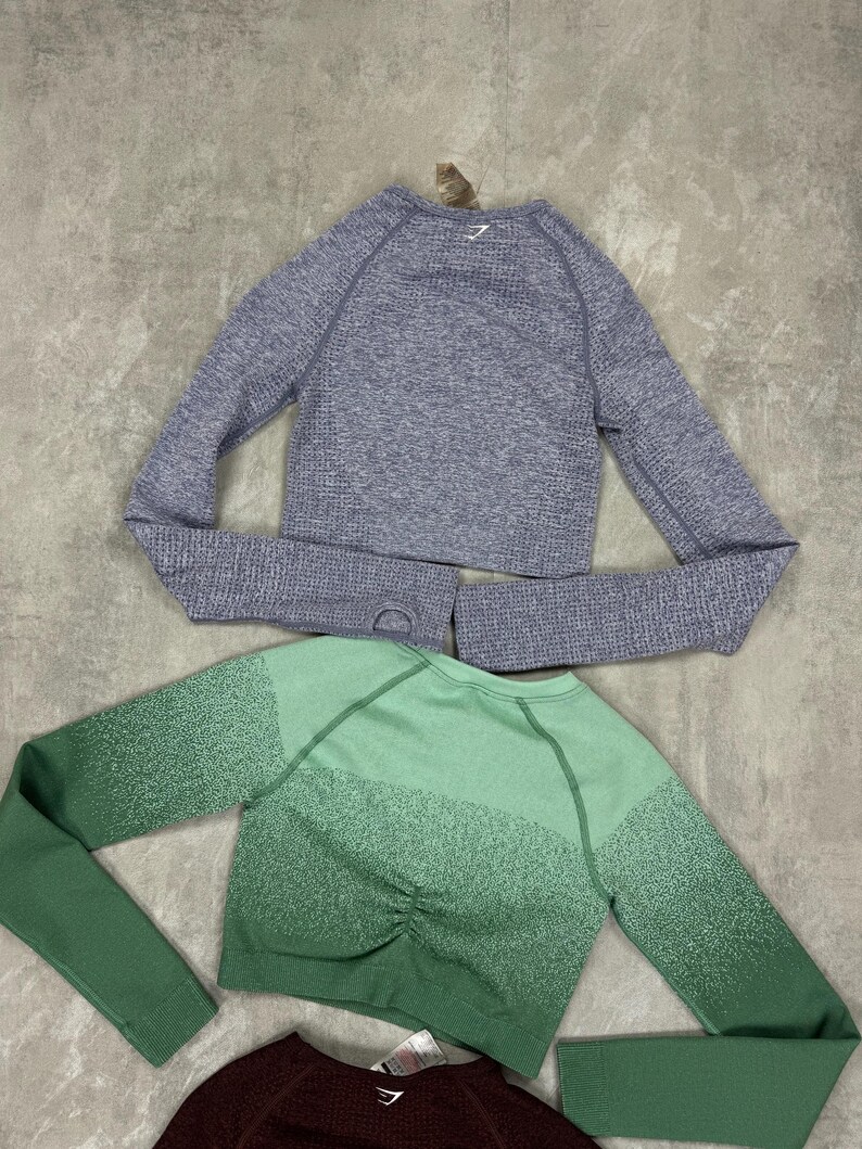 May include: Three long-sleeved crop tops in shades of gray, green, and burgundy. The gray top has a textured knit, while the green top features a gradient effect. Each top has a crew neck and thumb holes. The tops are designed for athletic wear.