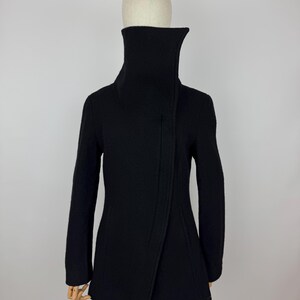 May include: A black coat with a high, asymmetrical collar. The coat has long sleeves and a wrap-style front. The coat is displayed on a mannequin against a plain white background. The coat's design is minimalist and modern.
