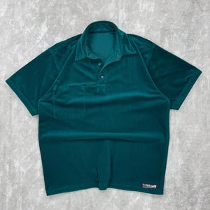May include: A teal green polo shirt with short sleeves. The shirt has a collared neckline with three buttons and a small pocket on the left chest. The fabric appears to be a soft, velvety material. The brand name "PARAMO" is printed on the bottom right.