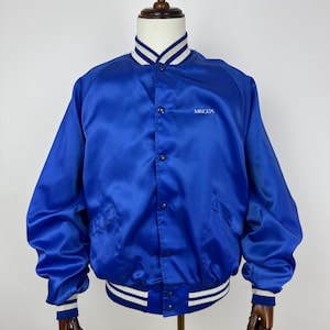 May include: A royal blue satin bomber jacket with white and navy striped trim on the collar, cuffs, and waistband. The jacket has a button-up front and two front pockets. The word "MINOLTA" is embroidered in white on the chest.