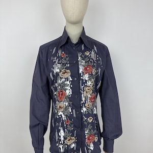 May include: A dark blue button-down shirt with long sleeves and a pointed collar. The shirt features floral embroidery with red, beige, and silver sequins down the front. The shirt is displayed on a mannequin.
