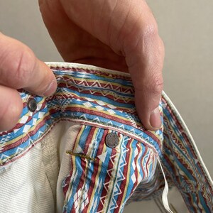 May include: White denim jeans with a colorful geometric pattern on the waistband. The pattern includes red, blue, yellow, and white.
