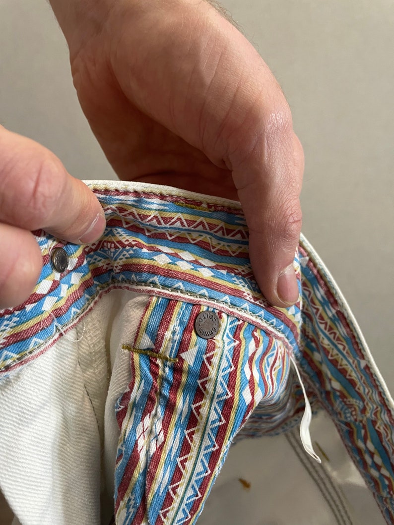 May include: White denim jeans with a colorful geometric pattern on the waistband. The pattern includes red, blue, yellow, and white.