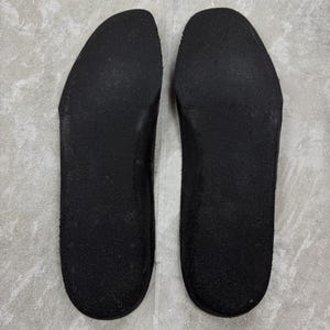 May include: Two black shoe insoles are displayed side by side. The insoles are made of a textured material and have a curved shape to fit the foot. The insoles are designed to be placed inside shoes for added comfort and support.