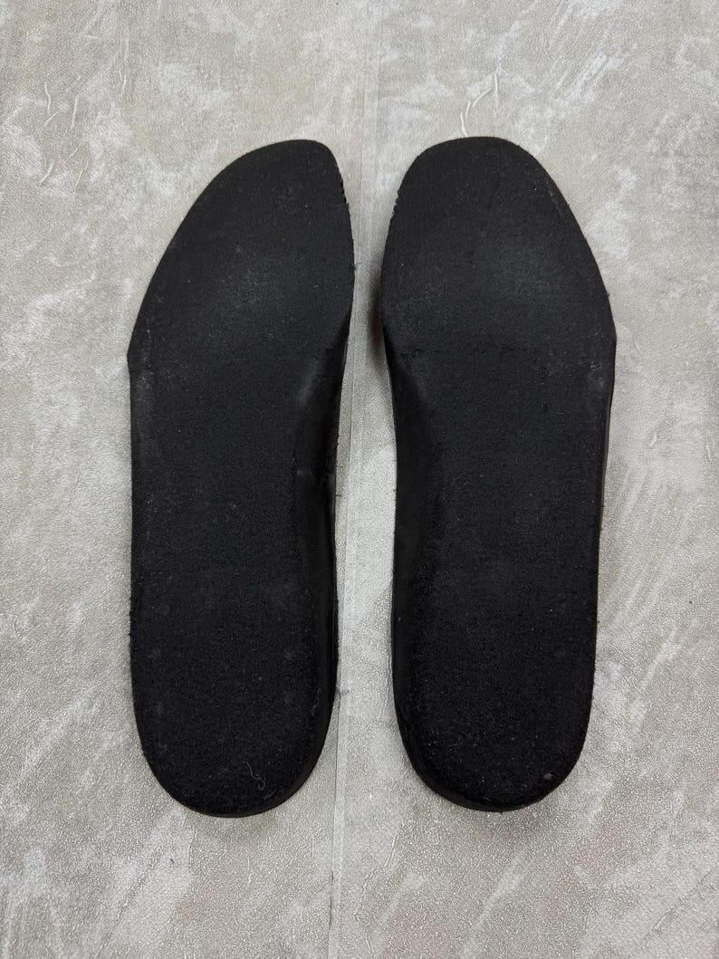 May include: Two black shoe insoles are displayed side by side. The insoles are made of a textured material and have a curved shape to fit the foot. The insoles are designed to be placed inside shoes for added comfort and support.