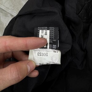 May include: A black garment interior featuring a white fabric care label with the text 100% POLYESTER and a secondary white tag displaying the number 16653, along with various laundry symbols and manufacturing details printed in black ink.