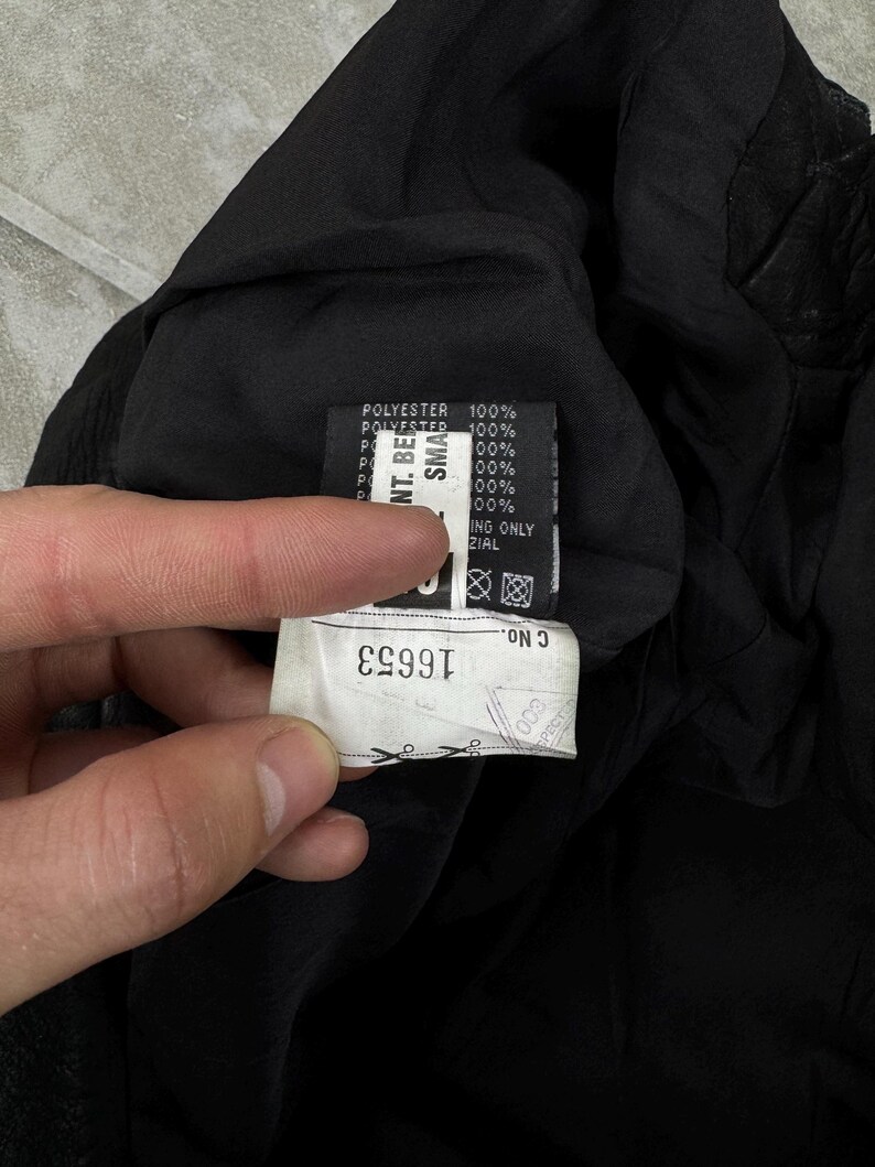 May include: A black garment interior featuring a white fabric care label with the text 100% POLYESTER and a secondary white tag displaying the number 16653, along with various laundry symbols and manufacturing details printed in black ink.