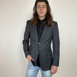 May include: A dark gray blazer with a single button closure. The blazer has a subtle texture and is worn over a black button-down shirt. The person is wearing light blue jeans.