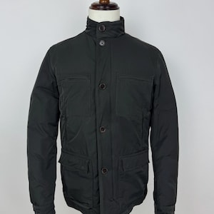 May include: A dark gray jacket with a high collar and multiple pockets. The jacket has a button-up front and is displayed on a mannequin. The jacket appears to be made of a smooth, possibly water-resistant material.