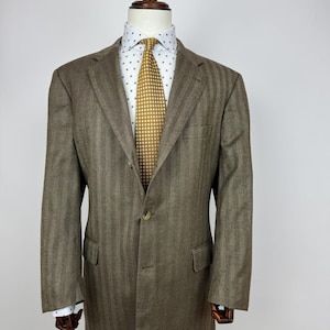 Puede incluir: Un blazer marrón con diseño de espiga exhibido en un maniquí. El blazer tiene cierre de dos botones, solapas con muescas y dos bolsillos delanteros. Se ve una camisa blanca con un pequeño estampado azul y una corbata dorada con un patrón repetitivo.