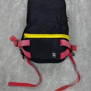 May include: Black backpack with yellow zipper and red straps. The backpack has a small logo on the front and is made of a lightweight material. The straps are adjustable and the bag appears to be designed for outdoor activities.