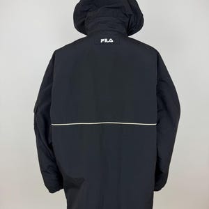 Puede incluir: Chaqueta negra con capucha y el logotipo blanco "FILA" en la parte superior de la espalda. La chaqueta tiene una raya horizontal blanca en la espalda y una capucha. La chaqueta est&aacute; hecha de un material ligero.