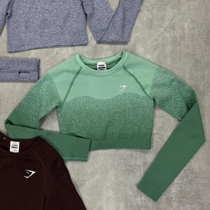 May include: Assortment of athletic wear in various colors. Includes a cropped green long-sleeve top, a gray long-sleeve top, and a maroon long-sleeve top. Each top features a white logo on the chest. A gray headband is also present.