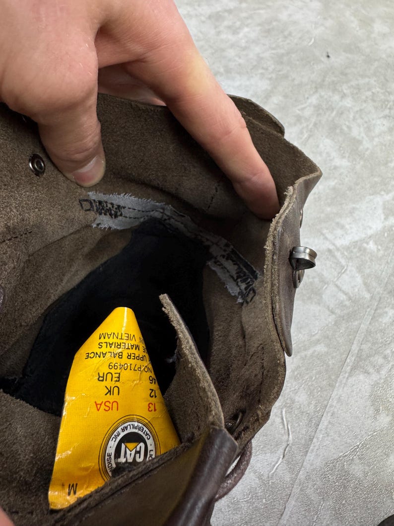 May include: Close-up of a brown leather boot interior. A yellow tag with the CAT logo and size information is visible. The boot has a worn appearance, with visible stitching and metal hardware.
